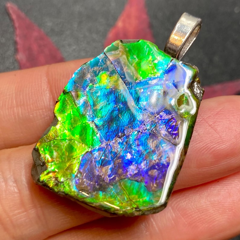 May include: A pendant featuring a raw, iridescent ammolite gemstone with a vibrant play of color, displaying intense shades of neon green, electric blue, and deep violet. The stone has a jagged, organic shape and is set with a silver-toned metal bail.