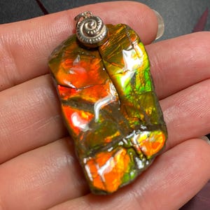 May include: A rectangular pendant featuring a polished piece of iridescent ammolite with vibrant orange, red, and green color play across its fractured surface. The stone is topped with a silver-toned metal bail featuring a spiral design.