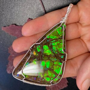 May include: A triangular pendant featuring a polished ammolite gemstone with vibrant green and iridescent flashes set in a silver wire-wrapped frame. The stone displays a mosaic-like pattern of dark brown and bright neon green segments.