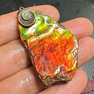 May include: A pendant featuring a polished, irregular piece of iridescent ammolite gemstone with vibrant red, orange, and green flashes. The stone is topped with a circular, silver-toned metal bail decorated with a raised spiral pattern.