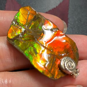 May include: A rectangular pendant featuring a polished piece of iridescent ammolite gemstone with vibrant orange, red, and green flashes. The stone has a natural cracked texture and is set with a small, silver-toned spiral metal bail at the bottom.