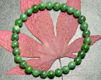May include: A circular bracelet composed of numerous small, smooth, spherical green beads. The beads exhibit varying shades of translucent and opaque green, arranged in a continuous loop. The bracelet is positioned over a large, dried red leaf.