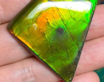 May include: A polished, iridescent gemstone with a diamond-like shape. The stone displays a vibrant array of colors, including green, gold, and orange, with visible internal fractures. The gemstone is held in a hand.