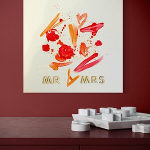 Könnte beinhalten: Eine weiße Leinwand mit abstrakten roten und orangen Farbstichen und Pinselstrichen. Die Worte "MR & MRS" sind in goldenen Buchstaben am unteren Rand der Leinwand geschrieben.