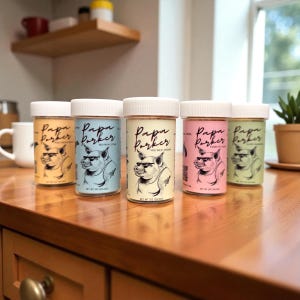 Puede incluir: Cinco frascos de especias con tapas blancas se exhiben sobre una superficie de madera. Cada frasco presenta una etiqueta de color pastel diferente con el texto "Papa Parker" y un cerdo de dibujos animados con gafas de sol. Los frascos están etiquetados como "Texas Country BBQ", "Big Papa's Steak", "Hog Wild Original", "Flavor Hot Chili" y "Hot Sauce".