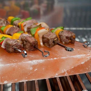 May include: Skewers of grilled meat and colorful vegetables rest on a large, rectangular pink Himalayan salt block. The skewers feature chunks of beef, bell peppers, onions, and tomatoes. The salt block sits on a metal grill.