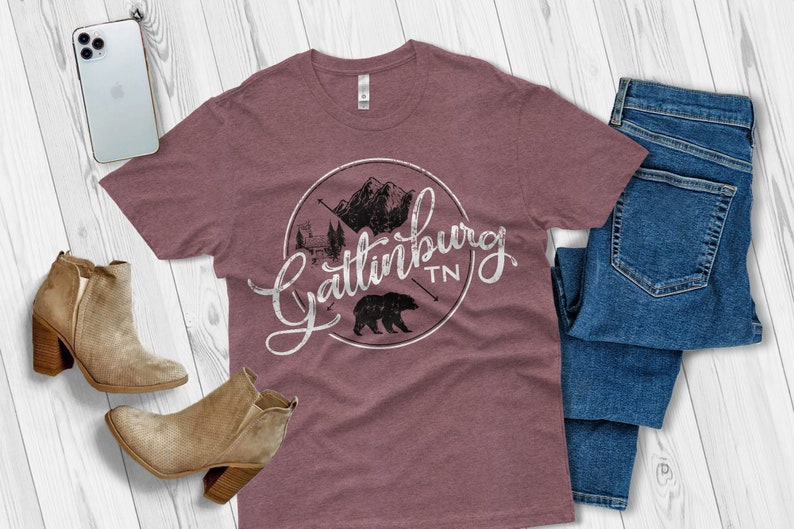 May include: A maroon t-shirt with a white graphic design of a circle with mountains, trees, a cabin, a bear, and the text "Gatlinburg TN".