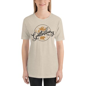 May include: A beige t-shirt with a black and brown graphic design that says "Gatlinburg TN" with a bear and mountains in the design.