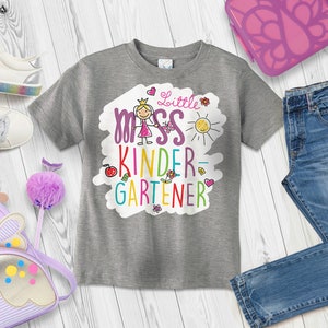 May include: A gray t-shirt with a colorful graphic that says "Little Miss Kinder-Gardener". The graphic features a stick figure girl wearing a crown and holding a butterfly. The shirt is laid out on a white wooden surface with other school supplies, including a purple backpack, a pair of jeans, a water bottle, and crayons.