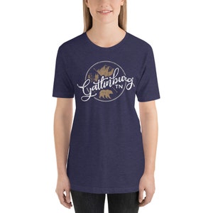 May include: A navy blue t-shirt with a white graphic design of a circle with the text "Gatlinburg TN" in a white script font. Inside the circle is a brown silhouette of a bear and a mountain range.