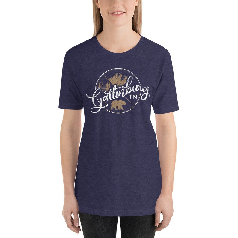 May include: A navy blue t-shirt with a white graphic design of a circle with the text "Gatlinburg TN" in a white script font. Inside the circle is a brown silhouette of a bear and a mountain range.
