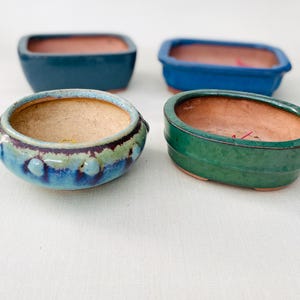 May include: Four ceramic bonsai pots in various shapes and colors. Two are rectangular with teal and blue glazes, one is a round bowl with a blue and green drip glaze, and one is an oval pot with a solid green glaze. All have terracotta interiors.