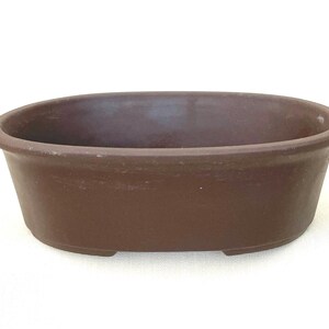 May include: An oval-shaped brown ceramic bonsai pot with a smooth, matte finish and a wide, rounded rim. The container features a deep basin and a solid base with small feet, designed for planting small trees or shrubs in a shallow soil depth.