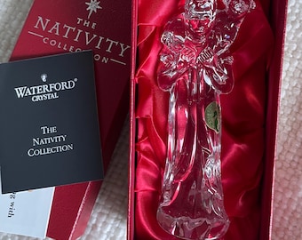 May include: A clear Waterford Crystal figurine from The Nativity Collection depicting a figure carrying a lamb on their shoulders. The figurine is housed in a red box with red satin lining and includes a black informational card with white text.