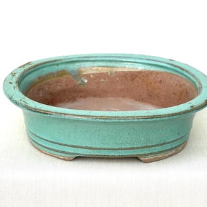 May include: An oval ceramic bonsai pot featuring a vibrant turquoise glaze on the exterior and a smooth, unglazed terracotta-colored interior. The vessel has a wide, flat rim, two horizontal incised lines around the base, and small feet.