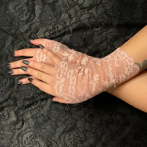 以下が含まれることがあります： 繊細な花柄のピンクのレースのフィンガーレスグローブのペア。グローブは透き通った生地で作られており、柔らかくフェミニンな印象です。