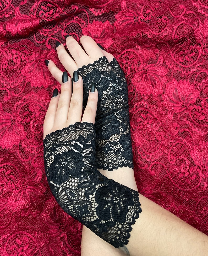 以下が含まれることがあります： 花柄の黒いレースのフィンガーレスグローブのペア。グローブは赤いレースの背景の上にあります。