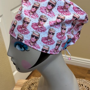 Op de afbeelding: Een wit en roze patroon scrub cap met een herhalend ontwerp van een man met een zonnebril die een kauwgom bel blaast. De cap heeft een trekkoord voor aanpassing en wordt weergegeven op een mannequin hoofd.