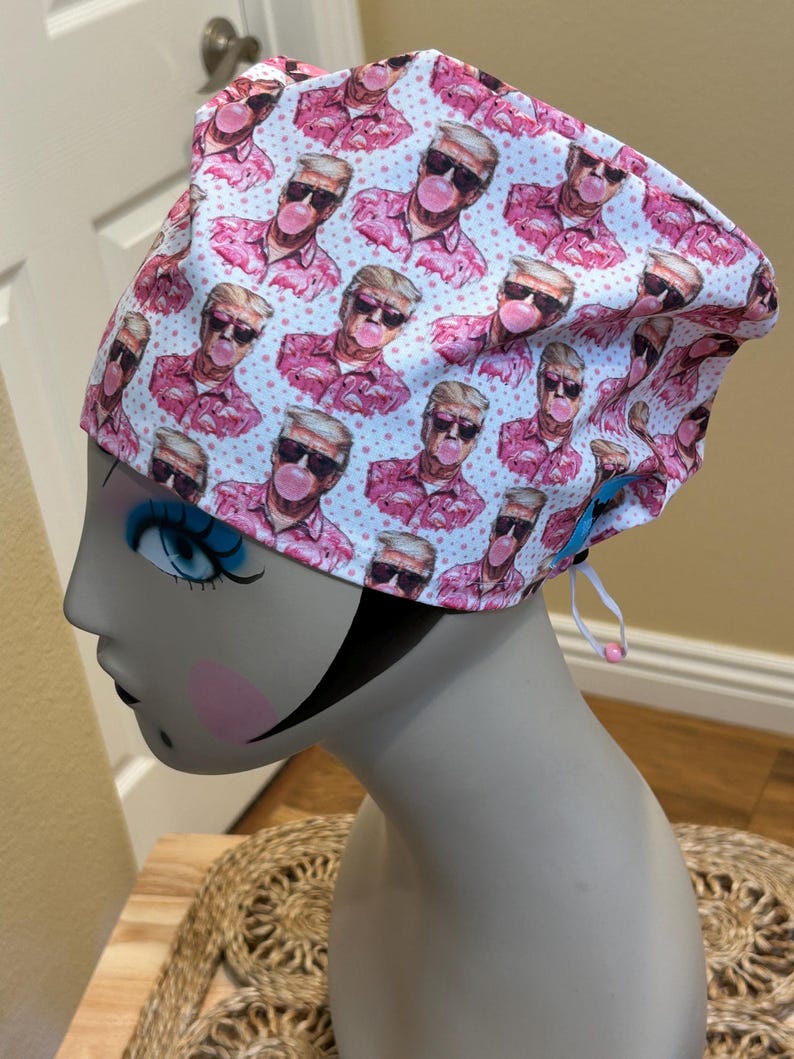 Op de afbeelding: Een wit en roze patroon scrub cap met een herhalend ontwerp van een man met een zonnebril die een kauwgom bel blaast. De cap heeft een trekkoord voor aanpassing en wordt weergegeven op een mannequin hoofd.