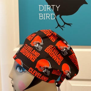 May include: A black and orange Cleveland Browns scrub cap. The cap features repeating patterns of the team's logo and the words "Cleveland Browns". The background includes a teal sign with a black bird silhouette and the words "Dirty Bird".
