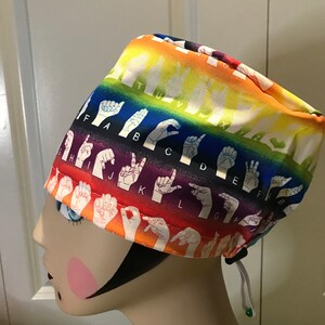 May include: A rainbow-colored surgical scrub hat with the letters of the American Sign Language alphabet printed on it. The hat has a drawstring.