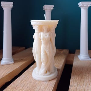 Puede incluir: Vela blanca en forma de columna con una figura femenina esculpida de pie sobre una columna. La vela est&aacute; rodeada de tres pilares blancos.