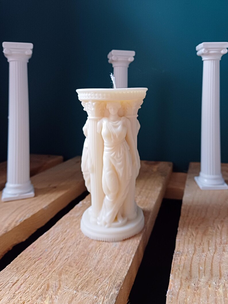 Puede incluir: Vela blanca en forma de columna con una figura femenina esculpida de pie sobre una columna. La vela est&aacute; rodeada de tres pilares blancos.