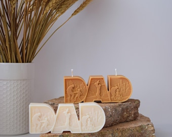 Puede incluir: Tres velas con la forma de la palabra "DAD" con escenas en relieve de un padre y un niño. Dos velas son de color naranja cálido y una es blanca. Las velas están sobre una superficie de piedra.