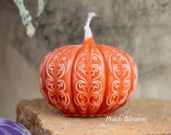 Peut inclure: Bougie orange en forme de citrouille avec des motifs décoratifs blancs. La bougie a une mèche blanche et est posée sur une surface en pierre. Le texte "Peach Blossom" est visible.