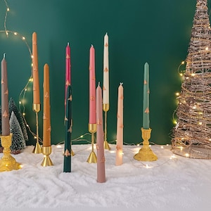 May include: A selection of colourful taper candles in shades of brown, pink, green, and white, decorated with gold Christmas tree designs. The candles are displayed in gold and brass candle holders, creating a festive holiday display.