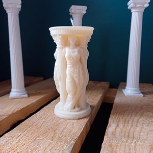 Puede incluir: Vela blanca con forma de columna con tres figuras de pie en la parte superior. La vela est&aacute; sobre una superficie de madera con dos columnas blancas a cada lado.