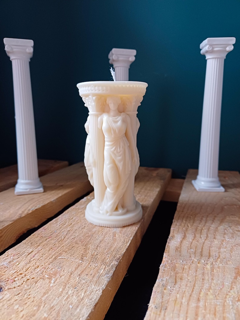 Puede incluir: Vela blanca con forma de columna con tres figuras de pie en la parte superior. La vela est&aacute; sobre una superficie de madera con dos columnas blancas a cada lado.
