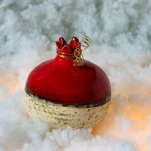 May include: A red and white ceramic pomegranate ornament with a gold wire detail. The ornament is sitting on a white fluffy surface.