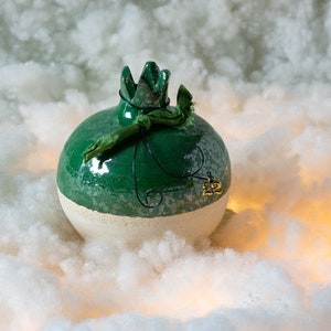 May include: A green and white ceramic pomegranate with a green ribbon tied around the top. The pomegranate has a gold charm with the number 22 on it.