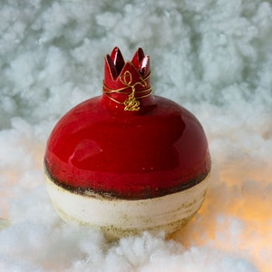 May include: A red and white ceramic pomegranate with a gold crown and the number 22 on top. The pomegranate is sitting on a white fluffy surface.