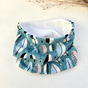 Op de afbeelding: Een teal en wit patroon nekwarmer met een zachte witte binnenkant. Het patroon bevat zwarte, witte en roze veervormige vormen. De nekwarmer is dubbelgevouwen, waardoor de binnenkant zichtbaar is.