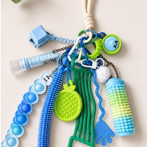 May include: A sensory keyring featuring a collection of blue and green fidget toys, including a gradient pop-it strip, a spiky tube, a textured pineapple charm, a green noodle pull, a blue hand shape, a spiky roller, and a small white pom-pom.