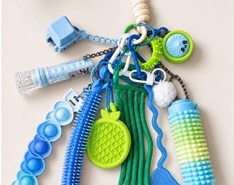 May include: A sensory keyring featuring a collection of blue and green fidget toys, including a gradient pop-it strip, a spiky tube, a textured pineapple charm, a green noodle pull, a blue hand shape, a spiky roller, and a small white pom-pom.