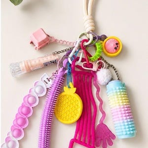 May include: A multi-coloured sensory keyring featuring a variety of tactile items including a pink pop-it strip, a purple spiky noodle, a yellow pineapple charm, a magenta ladder-shaped piece, a small hand-shaped charm, and a pastel rainbow roller.