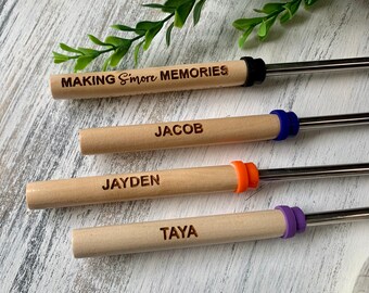 May include: Four personalized s'mores roasting sticks with wooden handles. The handles are engraved with names and the phrase "MAKING S'more MEMORIES." Each stick has a different colored silicone ring: black, blue, orange, and purple.