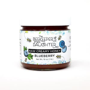May include: A glass jar of The Beekeeper's Daughter raw creamy blueberry honey. The jar has a white lid and a colorful label with illustrations of bees, blueberries, and flowers. The label reads "The Beekeeper's Daughter Raw Creamy Honey Blueberry Net Wt. 16 oz (1 lb.)"