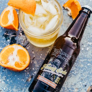 May include: A brown glass bottle of The Beekeeper's Daughter Honey Soda, Honey Bee Orange flavor, on a blue wooden surface with orange slices and a glass of ice water.