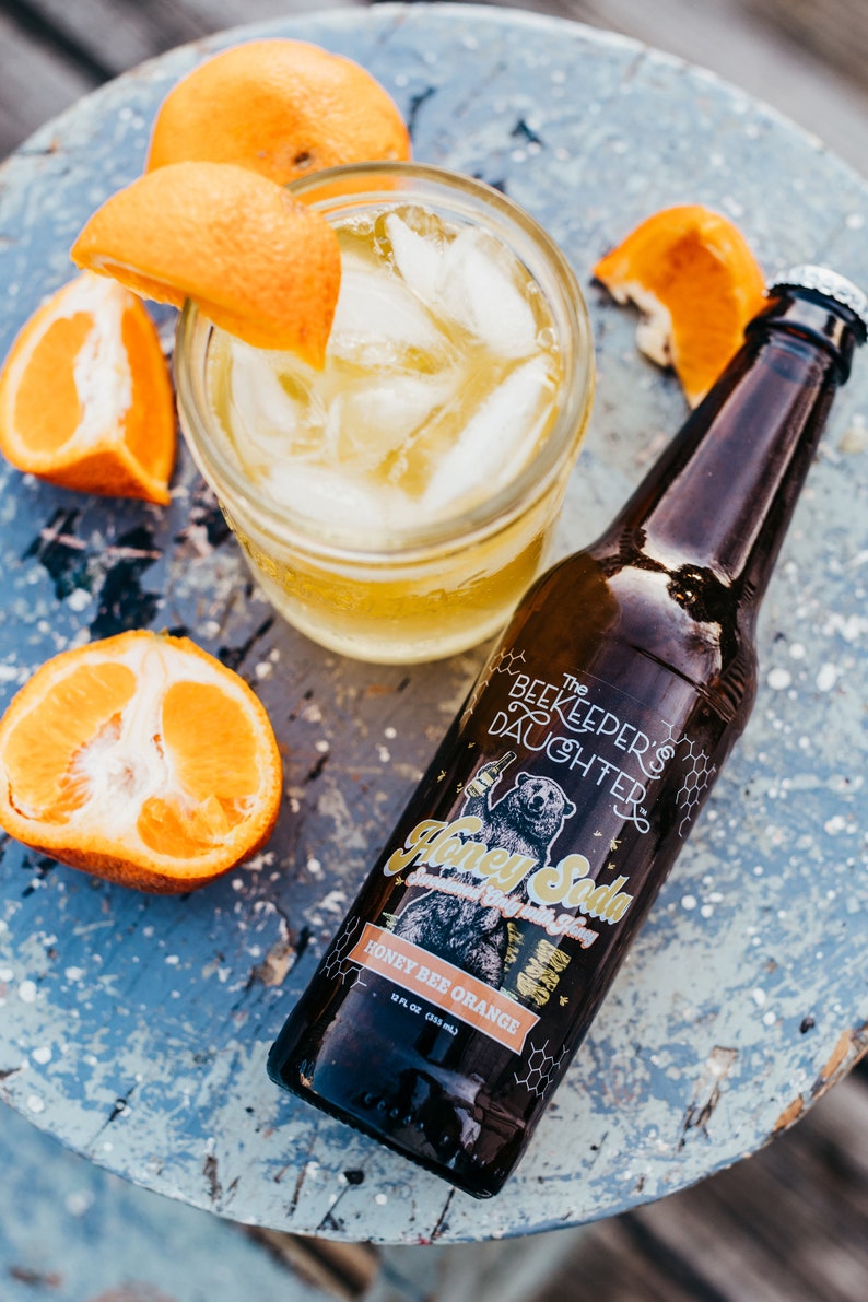 May include: A brown glass bottle of The Beekeeper's Daughter Honey Soda, Honey Bee Orange flavor, on a blue wooden surface with orange slices and a glass of ice water.