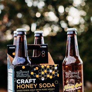 May include: Four-pack of 12-ounce glass bottles of The Beekeeper's Daughter Craft Honey Soda, Honey Bee Orange flavor. The bottles are brown glass with a silver cap. The packaging is black with a honeycomb pattern and a cartoon bee.
