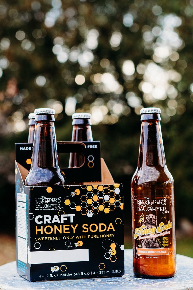 May include: Four-pack of 12-ounce glass bottles of The Beekeeper's Daughter Craft Honey Soda, Honey Bee Orange flavor. The bottles are brown glass with a silver cap. The packaging is black with a honeycomb pattern and a cartoon bee.
