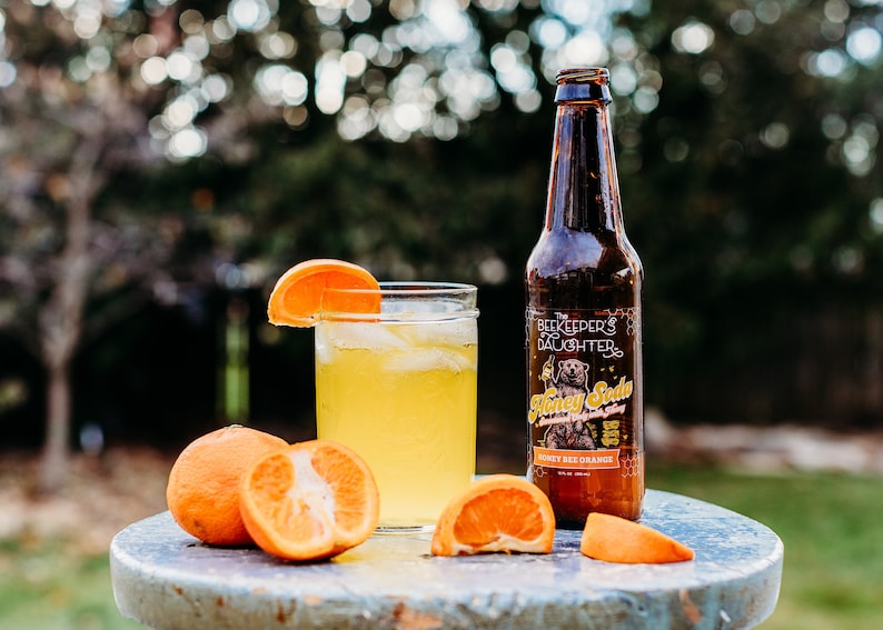May include: A glass of honey bee orange soda with ice and an orange slice garnish, next to a bottle of The Beekeeper's Daughter Honey Soda, Honey Bee Orange, 12 fl oz, and two orange halves on a blue table.