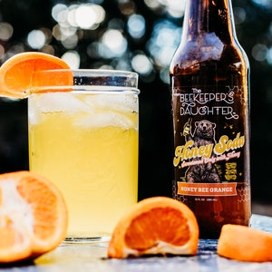 May include: A glass of honey bee orange soda with ice and an orange slice garnish. The soda is in a clear glass jar. A bottle of The Beekeeper's Daughter Honey Bee Orange Soda is in the background. The bottle is brown glass with a white label and a cartoon bear.