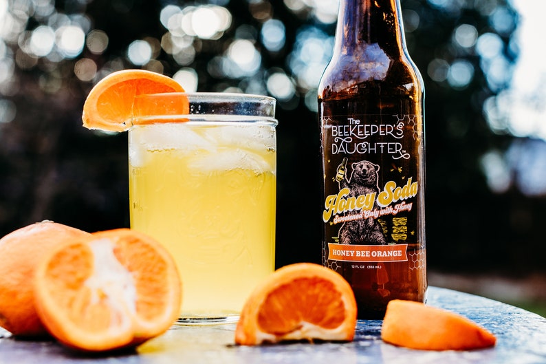 May include: A glass of honey bee orange soda with ice and an orange slice garnish. The soda is in a clear glass jar. A bottle of The Beekeeper's Daughter Honey Bee Orange Soda is in the background. The bottle is brown glass with a white label and a cartoon bear.