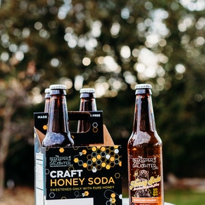May include: A six-pack of The Beekeeper's Daughter Craft Honey Soda in glass bottles. The packaging is black with yellow honeycomb designs and the text "Craft Honey Soda" and "Sweetened only with pure honey". One bottle is visible with the text "Honey Bee Orange".