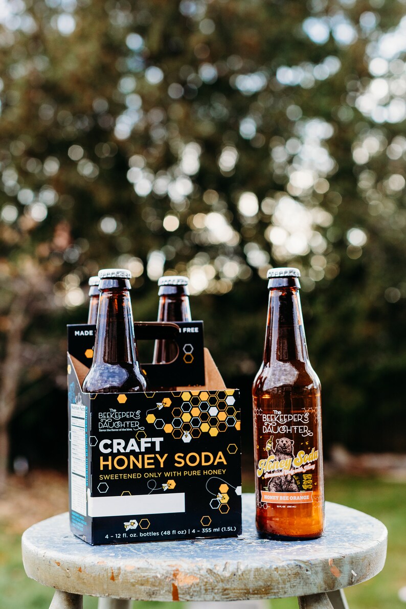 May include: A six-pack of The Beekeeper's Daughter Craft Honey Soda in glass bottles. The packaging is black with yellow honeycomb designs and the text "Craft Honey Soda" and "Sweetened only with pure honey". One bottle is visible with the text "Honey Bee Orange".